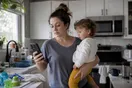 A parent stands in a kitchen holding a toddler on one hip while scrolling a phone with the other hand, waiting for a page to load.