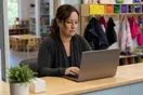 A daycare director sits at a small front-desk computer scrolling a slow-loading page on a laptop while cubbies and coat hooks line the wall behind her, in a natural documentary style.