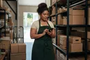 A small business owner stands in a stockroom checking a delivery company's website on a phone, boxes and a hand truck in the background, natural daylight, documentary style.