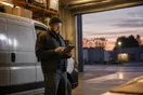A delivery business owner stands beside a parked van in a warehouse loading bay checking a tablet at dusk, in a natural documentary style.