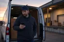 A delivery company owner standing beside his cargo van at a loading dock at dusk, reading an email on his phone.