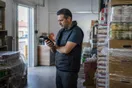 A small shop owner stands in a stockroom checking a phone while comparing last-mile delivery companies, photographed in a natural documentary style.