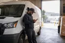 A delivery company owner checks his phone next to a parked cargo van in a warehouse loading bay, waiting for a page to load, in natural documentary light.