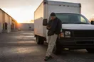 A delivery company owner stands beside a parked box van reviewing a slow-loading page on his phone at dawn, in a natural documentary style.