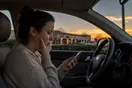 A woman sits in her parked car outside a strip mall at dusk, holding her jaw and scrolling her phone for a dentist who can see her soon.