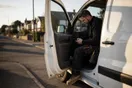 An electrician sits in his parked service van between calls, checking his phone on a quiet suburban street in late afternoon light.