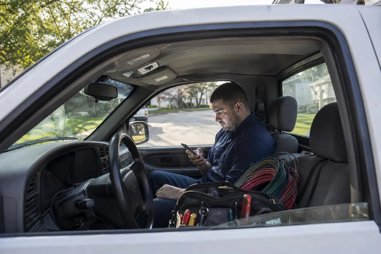 An electrician sits in the driver's seat of his parked service truck, checking a slow-loading page on his phone while coils of wire and a tool bag rest on the passenger seat, in a natural documentary style.