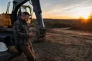 A contractor stands beside a parked excavator at the edge of a cleared lot at sunrise, checking messages on his phone before the crew arrives.