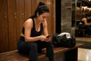 A person sits on a locker room bench in workout clothes scrolling a phone before a class, photographed in a natural documentary style.