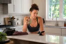 A woman scrolls gym websites on her phone at her kitchen counter early in the morning, coffee beside her, comparing intro offers before work.