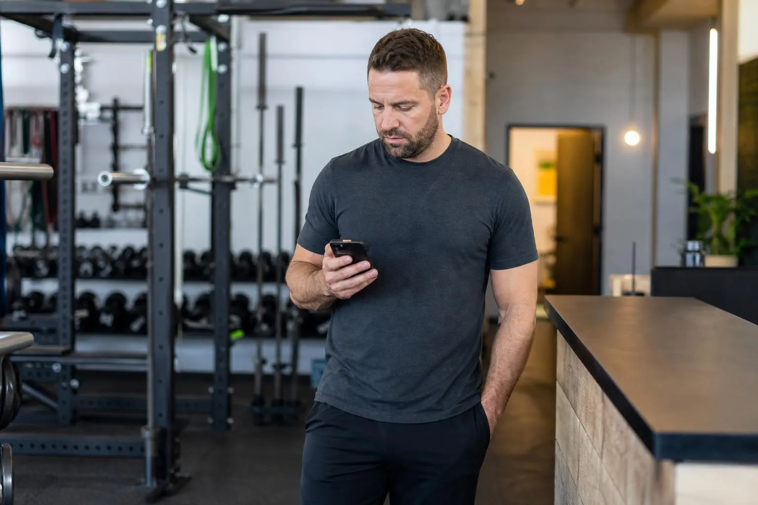 A gym owner stands near the front desk scrolling a slow-loading website on his phone while free weights and a chalk-dusted rack sit blurred behind him, in a natural documentary style.
