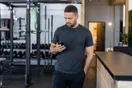 A gym owner stands near the front desk scrolling a slow-loading website on his phone while free weights and a chalk-dusted rack sit blurred behind him, in a natural documentary style.
