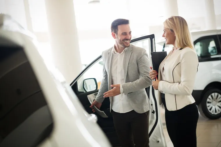 A dealership manager reviewing paperwork with a new sales hire at a desk on the showroom floor, in a documentary style.