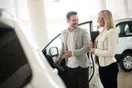 A dealership manager reviewing paperwork with a new sales hire at a desk on the showroom floor, in a documentary style.
