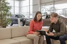 A car rental employee inspecting and cleaning a vehicle on a lot while checking items on a clipboard, in a natural documentary style.