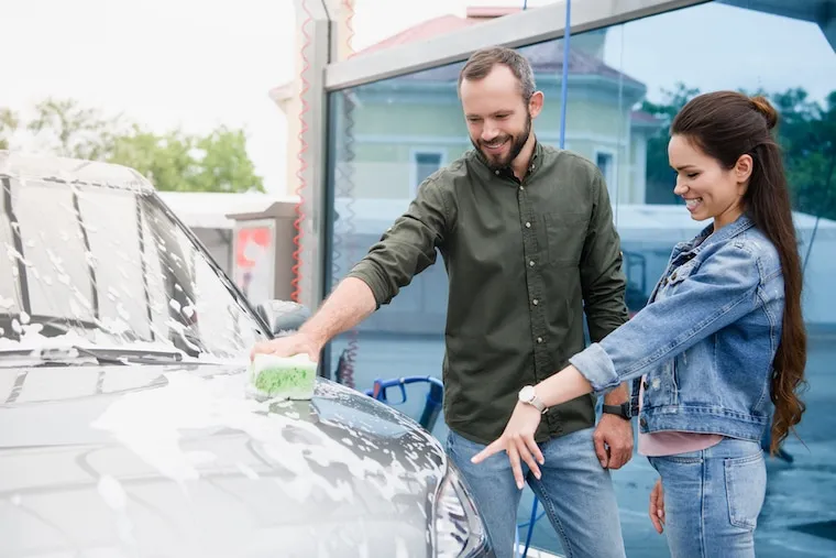 A car wash attendant in a branded shirt guiding a car onto the tunnel conveyor while a coworker preps the vehicle, in a natural documentary style.