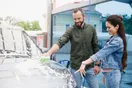 A car wash attendant in a branded shirt guiding a car onto the tunnel conveyor while a coworker preps the vehicle, in a natural documentary style.