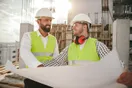 A construction company owner training a new crew member on a job site with tools and materials around them, in a natural documentary style.