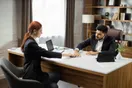 An attorney training a new paralegal at a shared desk with case files and a computer, natural documentary style.