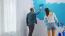 A lead painter showing a new hire how to cut in a wall edge on a job site, in a natural documentary style.