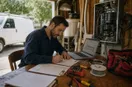 An electrician signing registration paperwork at a table with a laptop open, in a natural documentary style.