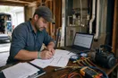 An HVAC technician signing registration paperwork at a table with a laptop open, in a natural documentary style.