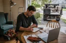 A junk removal operator at a kitchen table totaling startup costs with a calculator, notebook, and receipts, in a natural documentary style.