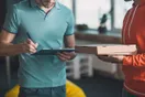 A courier business owner at a kitchen table with a laptop, a calculator, and a spreadsheet of startup costs, in a natural documentary style.