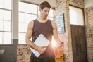 A gym owner working through a startup budget on a laptop and calculator at a bench inside a half-built gym, in a natural documentary style.