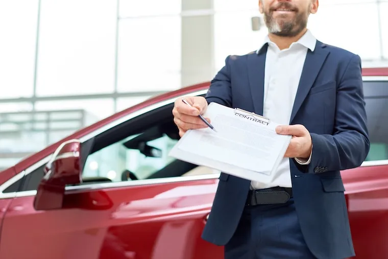 A car rental owner reviewing a fleet profit spreadsheet on a laptop with cars visible through a window, natural documentary style.