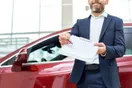 A car rental owner reviewing a fleet profit spreadsheet on a laptop with cars visible through a window, natural documentary style.