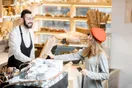 A baker handing a boxed order across a farmers market stall stacked with breads and pastries, in a natural documentary style.