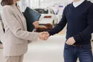 A rental agent handing car keys to a customer over a service counter with a fleet visible outside, documentary style.
