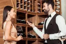 A tasting-room host pouring wine for two seated guests at a bar lined with bottles, in a natural documentary style.