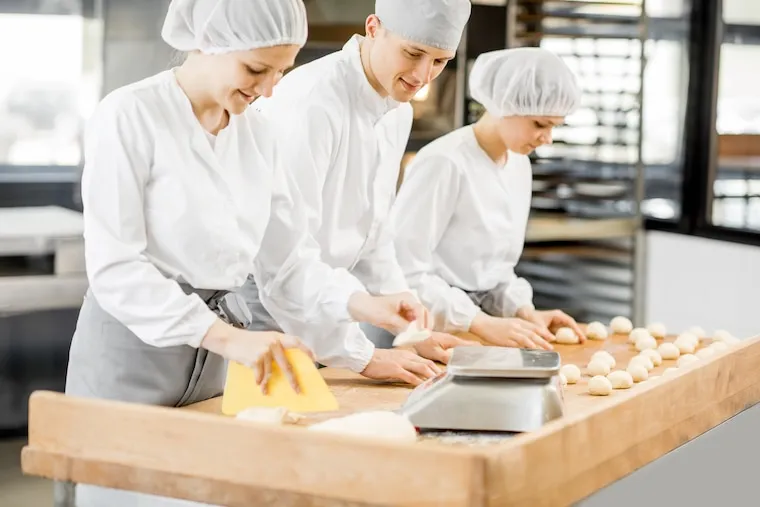 A baker and an assistant working side by side at a stainless prep table trays of loaves cooling behind them, in a natural documentary style.