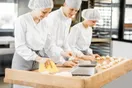A baker and an assistant working side by side at a stainless prep table trays of loaves cooling behind them, in a natural documentary style.
