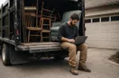 A junk-removal operator sitting on the open tailgate of a dump truck working on a laptop, with stacked wooden chairs and a teal armchair visible in the truck bed.