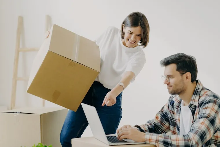 A mover reviewing a moving company website on a laptop at a kitchen table, boxes stacked in the background, natural documentary style.