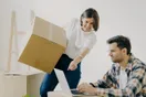 A mover reviewing a moving company website on a laptop at a kitchen table, boxes stacked in the background, natural documentary style.