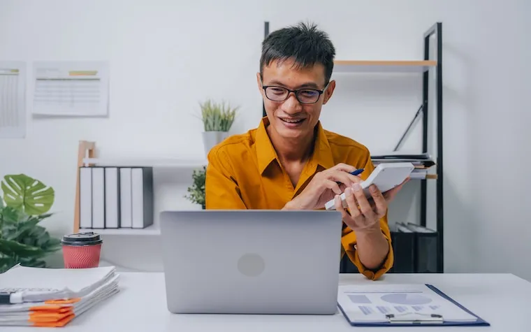 A smartphone showing a social media feed held in front of a desk with accounting paperwork, in a natural documentary style.