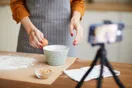 A baker photographing a decorated layer cake on a marble counter with a phone on a small tripod and window light, in a natural documentary style.