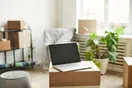 A mover recording a how-to packing tutorial on a tripod-mounted camera in a living room full of boxes, natural documentary style.