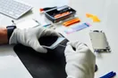 A technician recording a close-up micro-soldering repair on a phone board with a phone camera on a tripod, documentary style.
