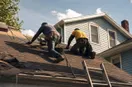 Two roofers laying fresh shingles on a residential roof on a sunny afternoon, shot from a low angle with sky and the neighbor's siding visible.