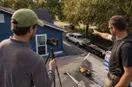 A phone tripod set up while a roofing contractor demonstrates equipment, in a natural documentary style.