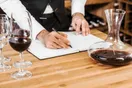 A winery owner reviewing federal permit paperwork at a desk with a laptop and a bottle of wine, in a natural documentary style.