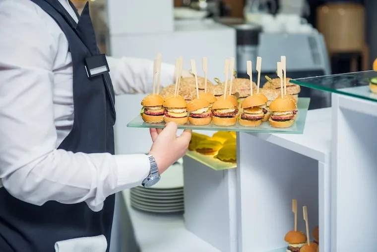 A catering team plating dishes on a stainless steel prep line at an event, in a natural documentary style.