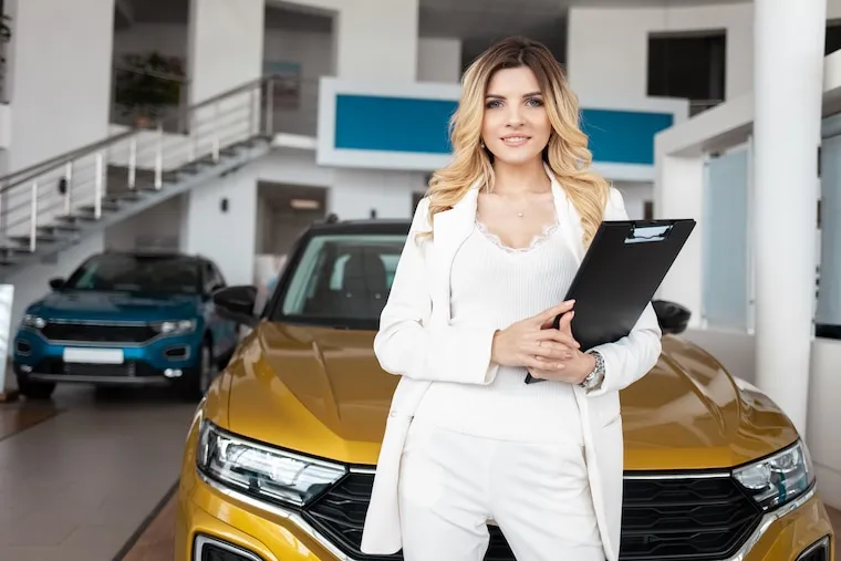 A dealership office desk with a laptop showing vehicle listings and a stack of deal folders, in a natural documentary style.