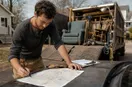 A junk removal operator marking up a paper city map on the hood of a truck, in a natural documentary style.