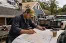 A roofing contractor marking up a paper city map on the hood of a truck, in a natural documentary style.